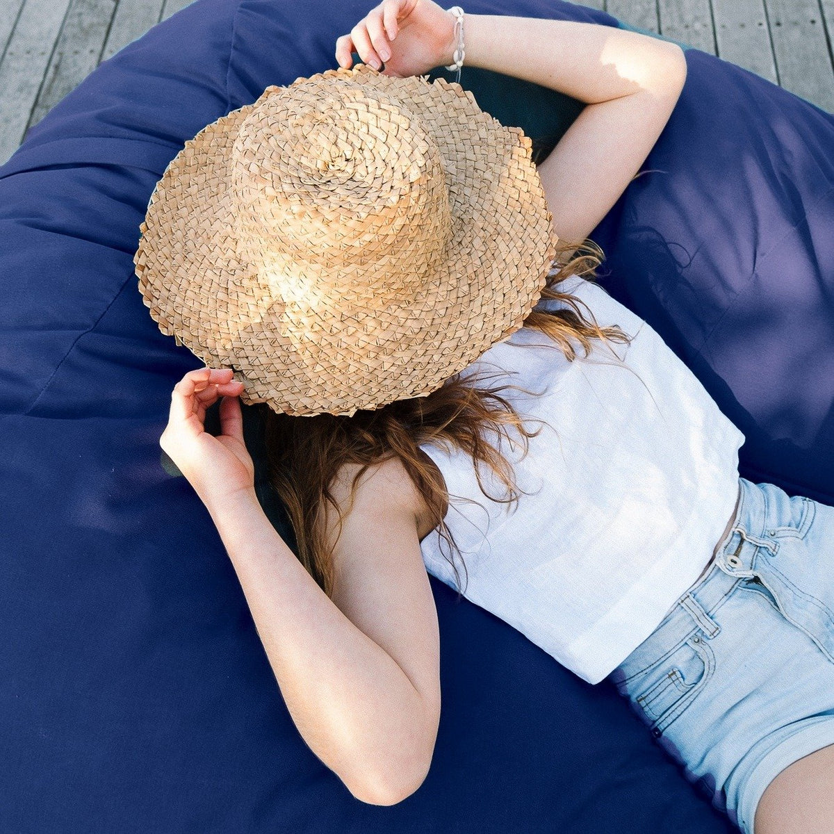 Girl in a straw hat lying on a Navy Shloomfy patio beanbag by the pool
